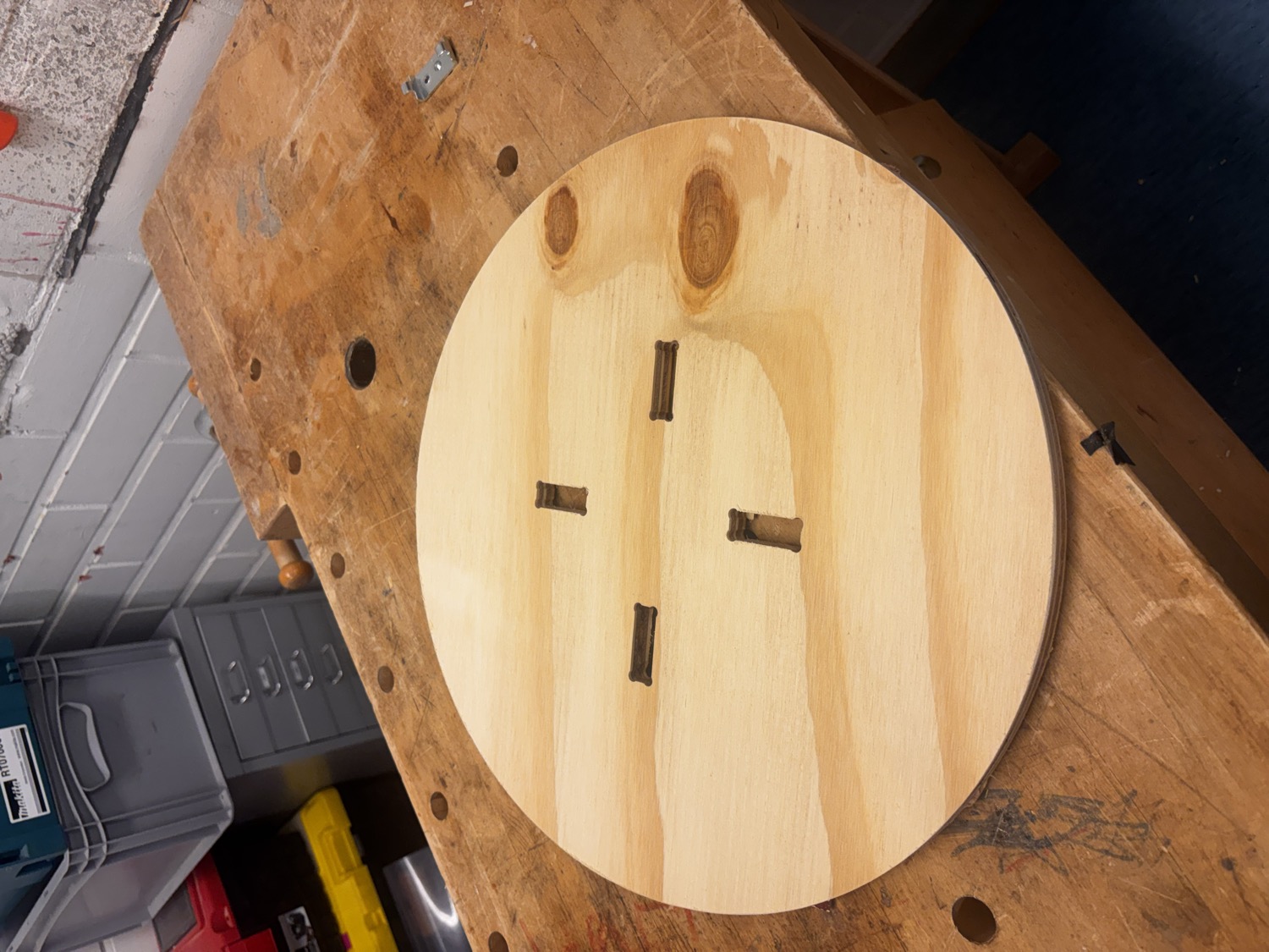 Round plywood table top resting on a workbench, showing the four through-cut finger-joint slots