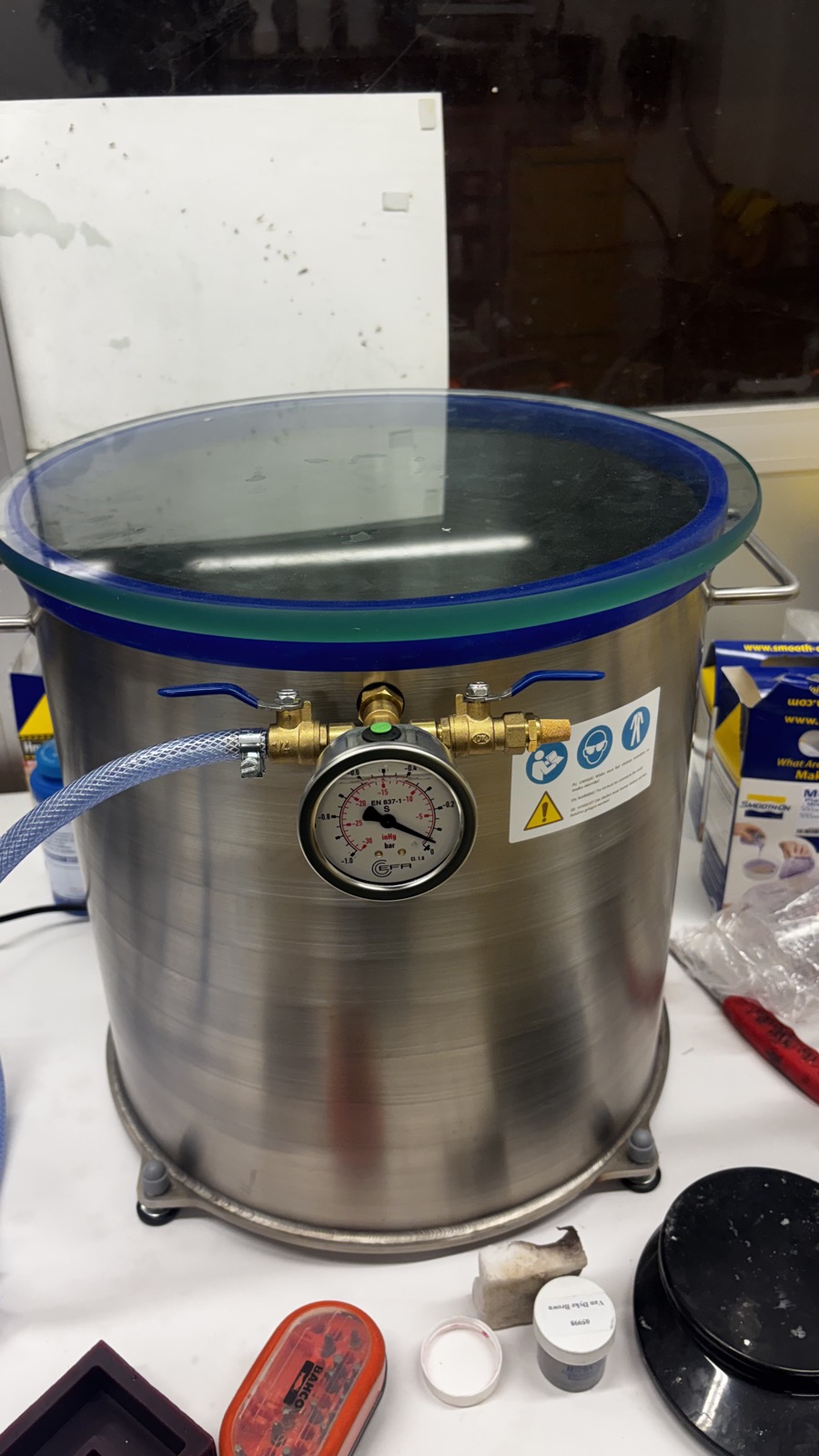 The stainless steel vacuum chamber with glass lid, brass valves and gauge on the casting room bench, hose attached