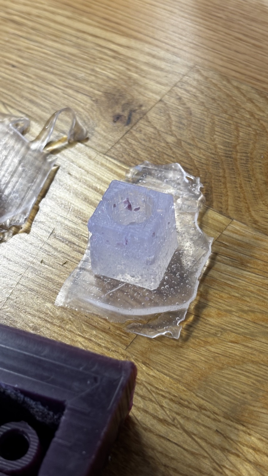 Close view of a demolded silicone mold cube with the knob cavity on top, sitting on its sheet of clear flash, flecks of purple wax embedded in the surface