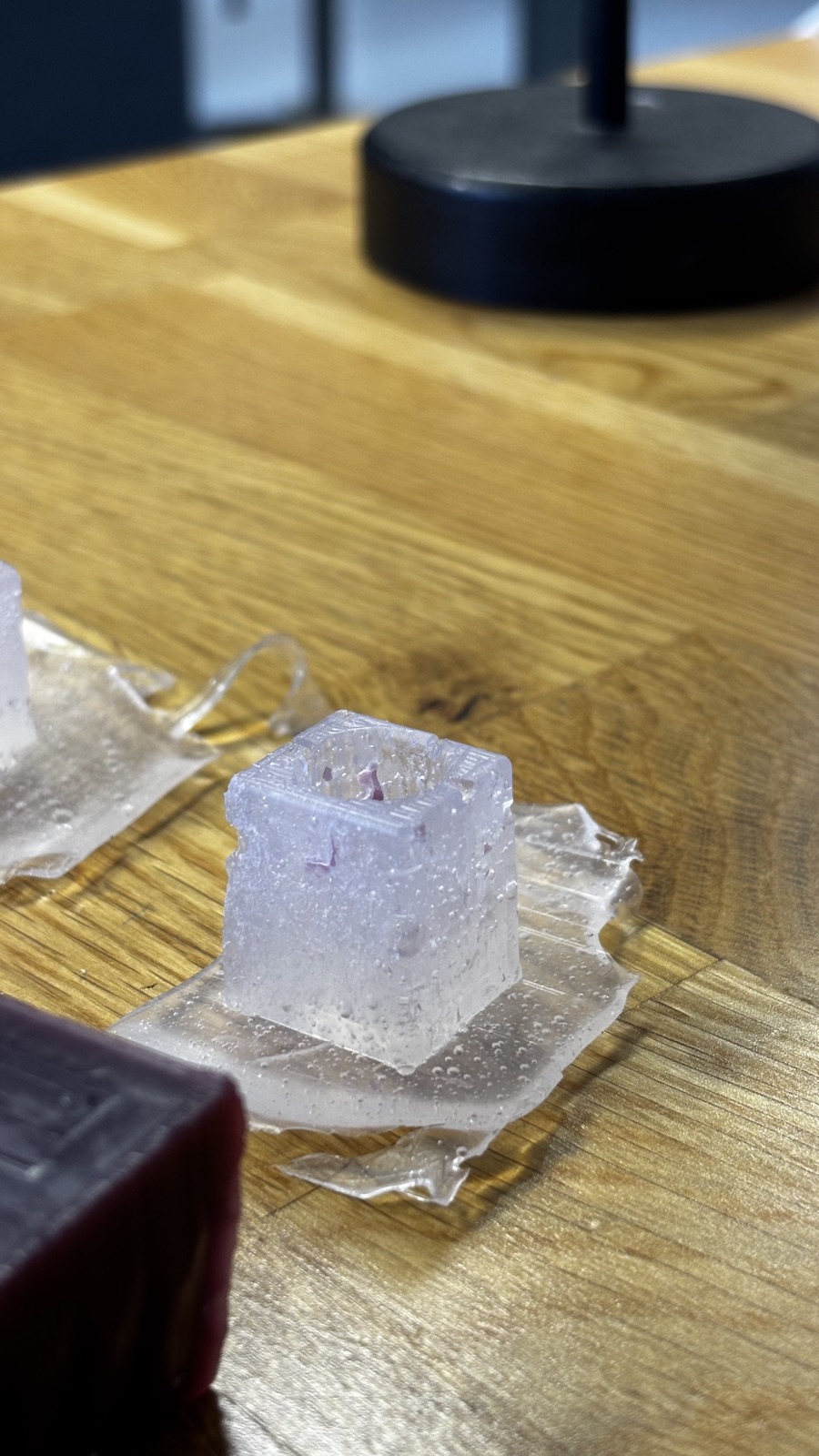 The second silicone mold cube on its flash sheet next to the edge of the wax block, the round knob cavity visible on top with small bubbles in the surrounding sheet