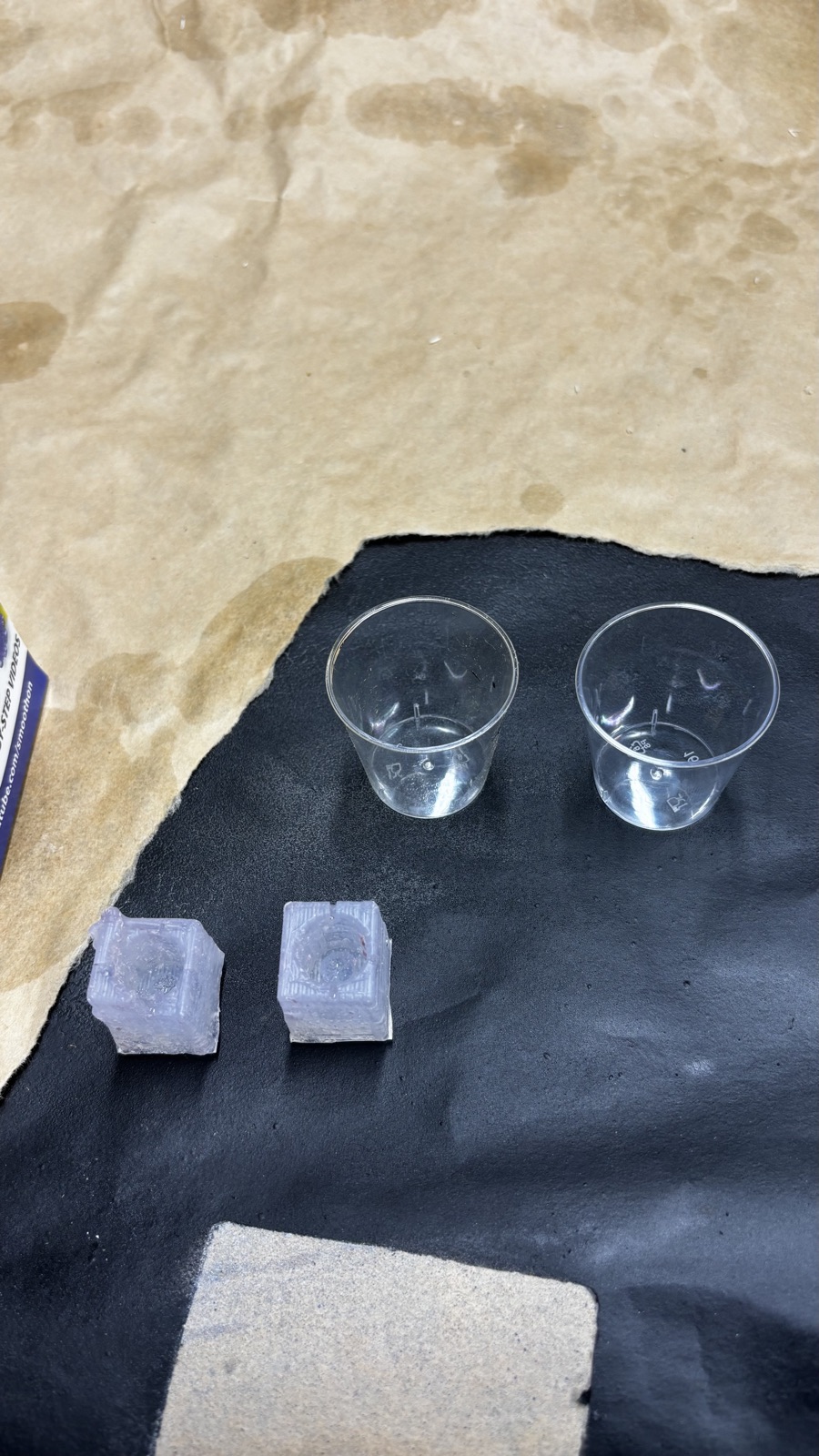 Two empty plastic mixing cups on the black mat with the two trimmed silicone molds below them