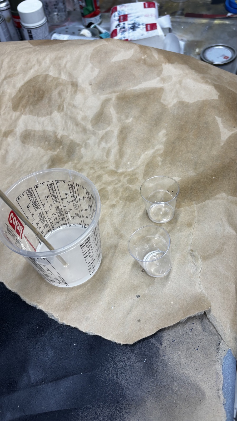 A graduated mixing cup with ratio markings holding the combined white resin and a wooden stir stick, the two emptied small cups beside it