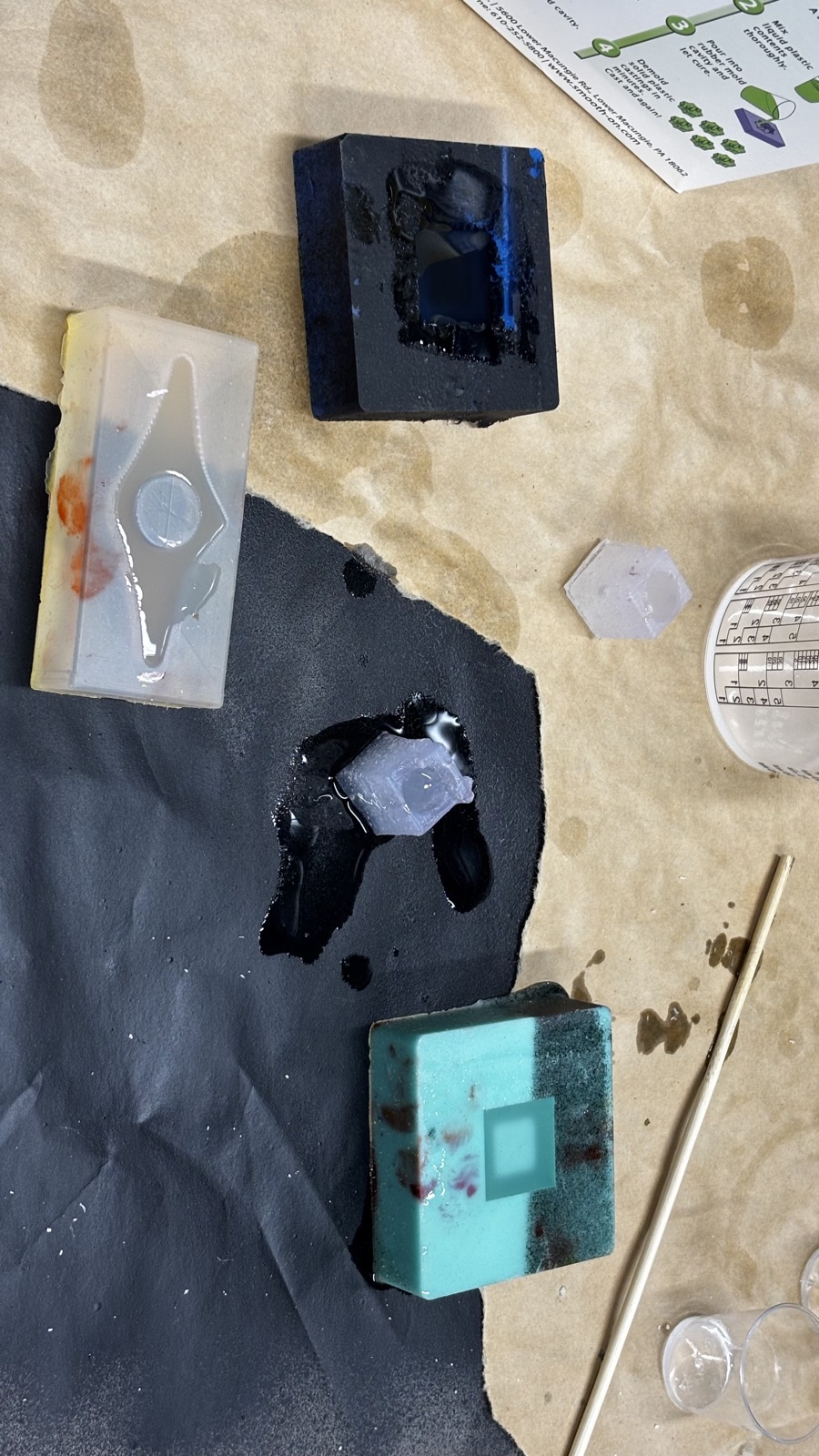 Found silicone molds on the bench holding leftover resin, a white mold with a pointed cavity, a dark mold, a teal mold with a square cavity, a small hexagonal piece and a mold cube sitting in a pool of spilled resin