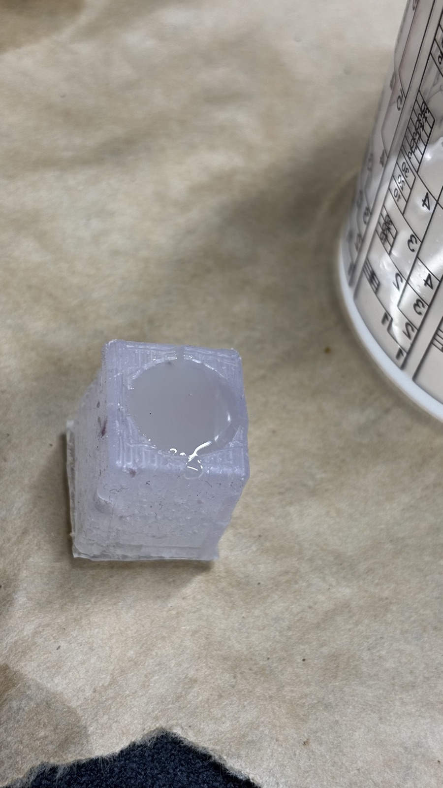 The trimmed silicone knob mold with its round cavity filled with white resin, a graduated mixing cup lying behind it