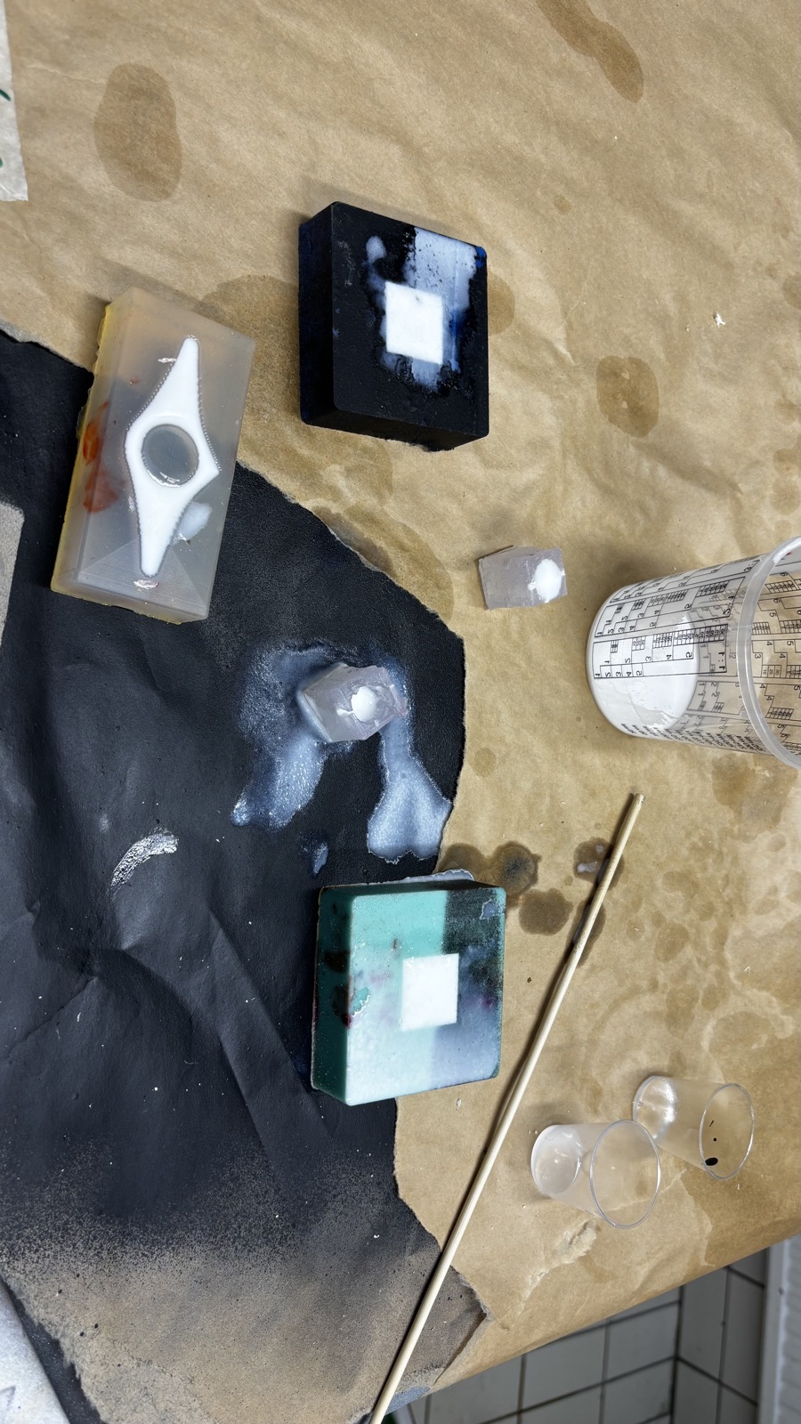 The bench after the cure, a dark blue block and a teal block each holding a white square of resin, a clear mold with a pointed white shape, two small silicone pieces with white resin and the emptied mixing cups on the brown paper