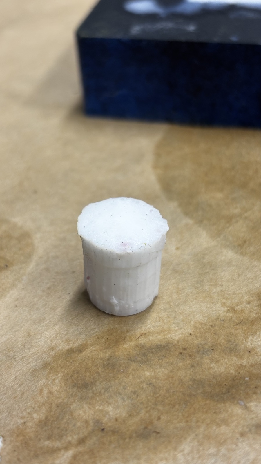 The demolded white knob standing on the brown paper, fine vertical ridges on its side and a rough bubbled top face, the dark blue mold block blurred behind it