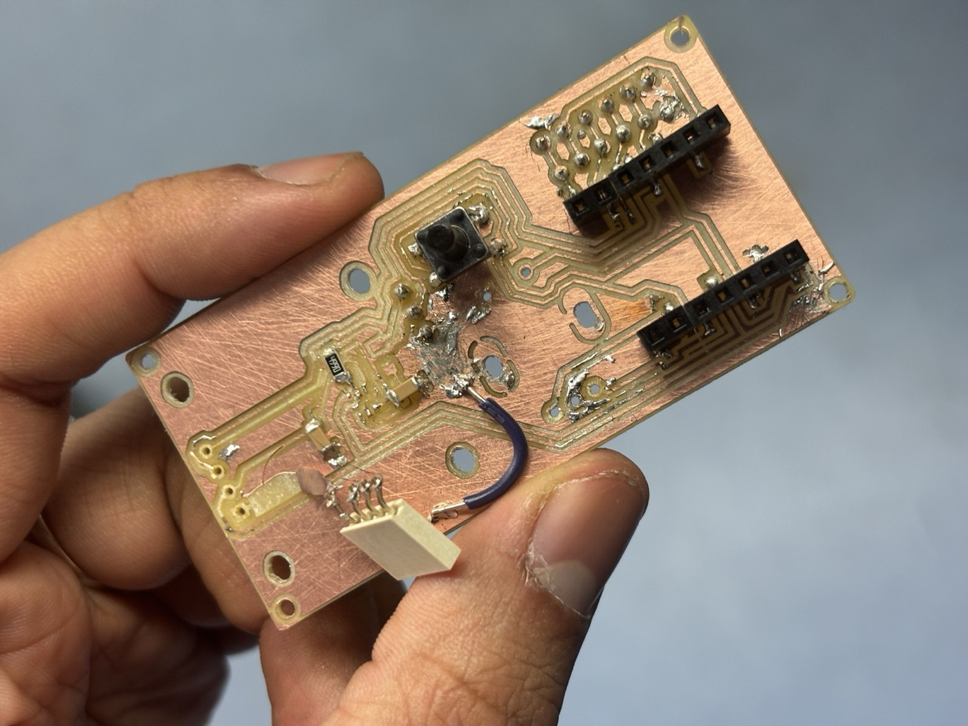 A milled and partially populated PCB held in a hand, showing solder bridges, jumper wires and patched traces from manual fixes