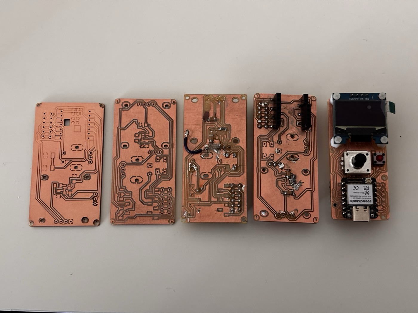 Six different milled PCB attempts laid out side by side on a workbench, showing different trace widths, isolation cuts and copper damage between iterations