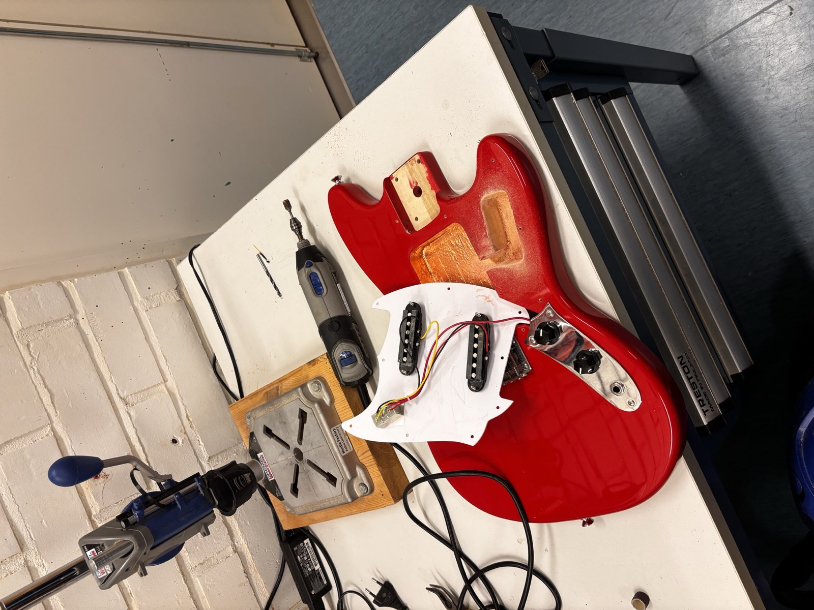 A red Squier Mustang body on a workbench next to a drill press and a rotary tool, with a freshly routed lighter-coloured pocket cut into the wood