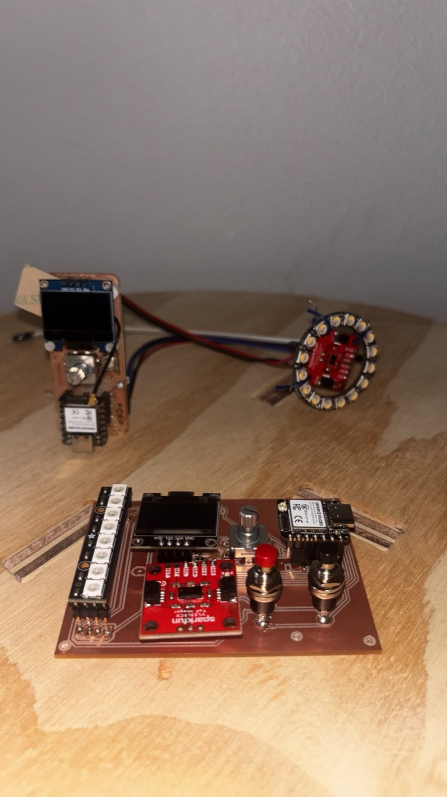 Two milled PCBs on a wooden surface: at the back, the guitar controller board with an OLED and an LED ring connected by wires; at the front, the new receiver board with an OLED, buttons and a XIAO ESP32-S3