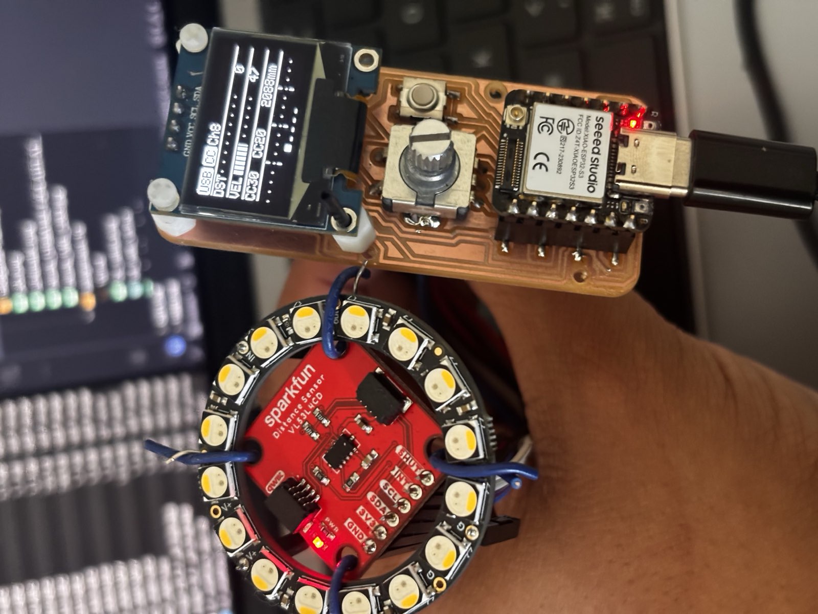 The guitar controller board powered over USB-C, its OLED showing live distance and CC values, next to a SparkFun time-of-flight sensor mounted inside an LED ring